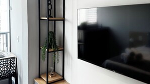 En moderne stue med et fladskærms-tv på væggen, træhylder, en lille plante og et spiseområde med bord og stole.