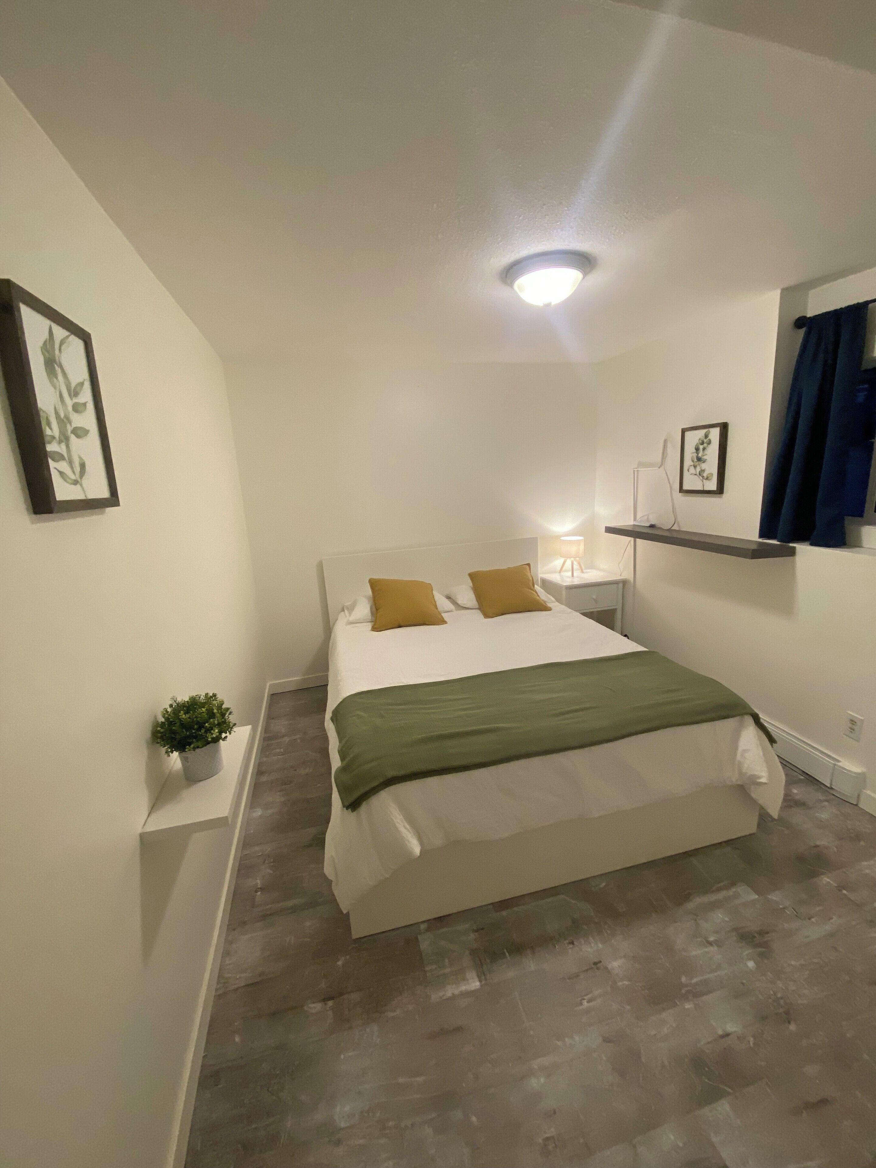 Un dormitorio con cama, mesita de noche, lámpara, estante con planta y luz de techo.
