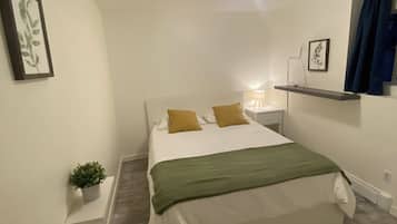 Un dormitorio con cama, mesita de noche, lámpara, estante con planta y luz de techo.