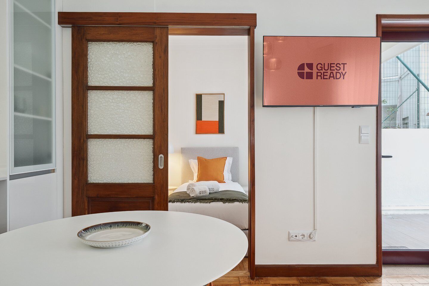 A hotel room with a wooden door, a white table with a bowl, a bed with orange pillows, and a 'GUEST READY' sign.