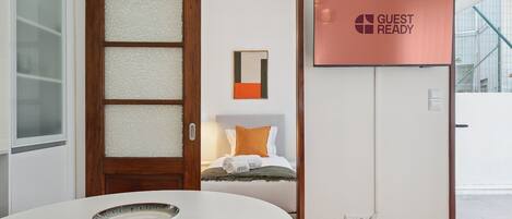 A hotel room with a wooden door, a white table with a bowl, a bed with orange pillows, and a 'GUEST READY' sign.