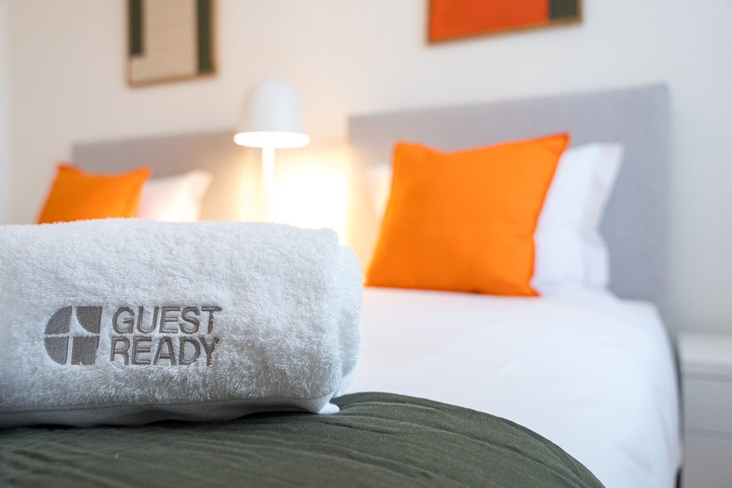 A hotel room with a bed, white bedding, an orange pillow, and a white towel with "GUEST READY" embroidered on it.