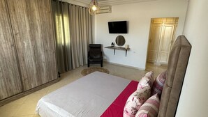 A spacious bedroom with a large bed, a flat-screen TV mounted on the wall, a wooden wardrobe, and a small round table with a green plant. - A pleasant ground floor 5 minutes' walk from the beach (Hergla)