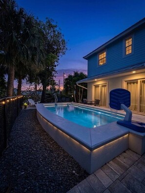 Una piscina con bordo curvo, una casa blu con garage e un albero sullo sfondo.
