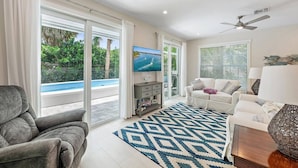 A modern living room with a pool view, a flat-screen TV, and a ceiling fan. - Movie Theater, Heated Pool, & Sauna - Luxe 4BR (West Palm Beach)
