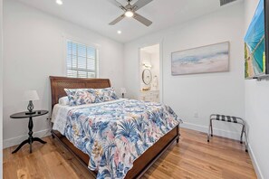 A bedroom with a wooden bed, a ceiling fan, a bedside table, a nightstand, a mirror, a painting, and a window with blinds.