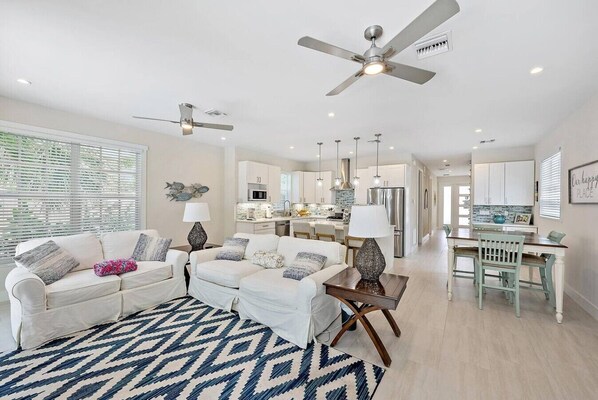 A modern living room with a sofa set, a patterned rug, ceiling fans, and a kitchen in the background.