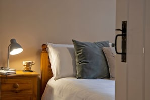 A neatly made bed with white and grey pillows, a wooden nightstand with a lamp, and a cup and book beside it.