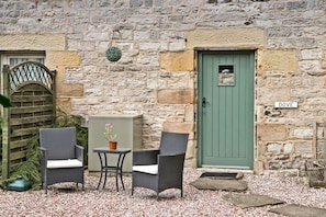 A stone building with a green door labeled "DOVE," two wicker chairs, a small table with a potted plant, and a wooden gate.