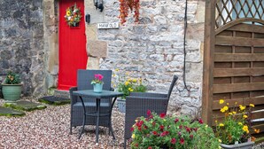 A stone building with a red door, a sign reading "MANIFOLD," outdoor wicker furniture, and various potted plants.