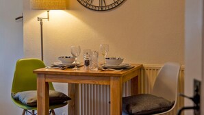 A dining area with a wooden table set for two, two chairs, a wall clock, and a floor lamp.