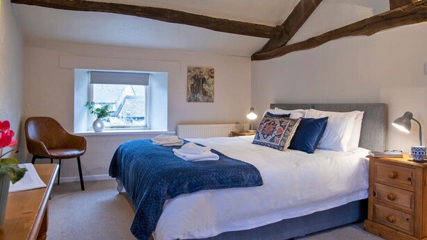 A bedroom with a large bed, a wooden chair, a nightstand, and a window with a view of a house.