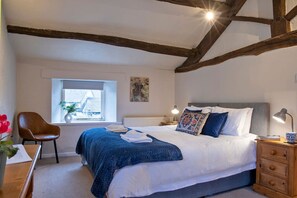 A bedroom with a large bed, a wooden chair, a nightstand, and a window with a view of a house. - Manifold Cottage - Central and Cosy (Hartington)