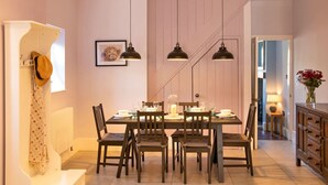 Ein Essbereich mit einem Holztisch, gedeckt für sechs Personen, umgeben von Stühlen. Über dem Tisch hängen Pendelleuchten, an der Wand hängt ein gerahmtes Bild und links befindet sich eine Heizung.