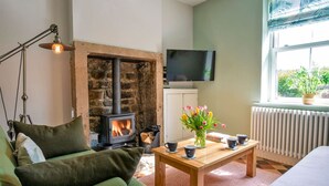 A cozy living room with a stone fireplace, a television mounted on the wall, and a window with a view of greenery. - Beech Cottage - Central Luxury (Derbyshire)