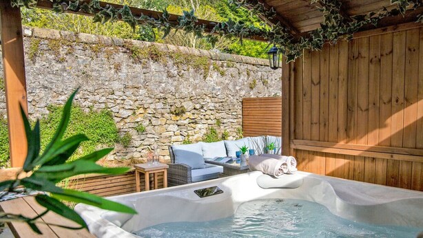A hot tub with a wooden deck, a stone wall, and a wooden pergola with greenery. - Beech Cottage - Central Luxury (Derbyshire)