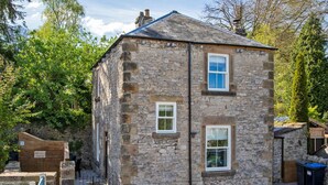 A stone building with two windows and a small door, surrounded by trees and a paved area. - Beech Cottage - Central Luxury (Derbyshire)