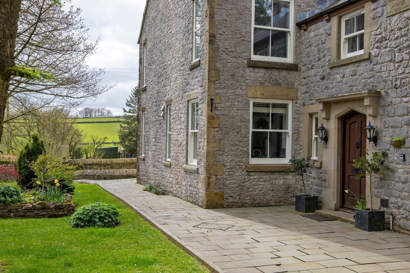 A stone house with a paved walkway, a garden with various plants, and a view of a green field in the background.