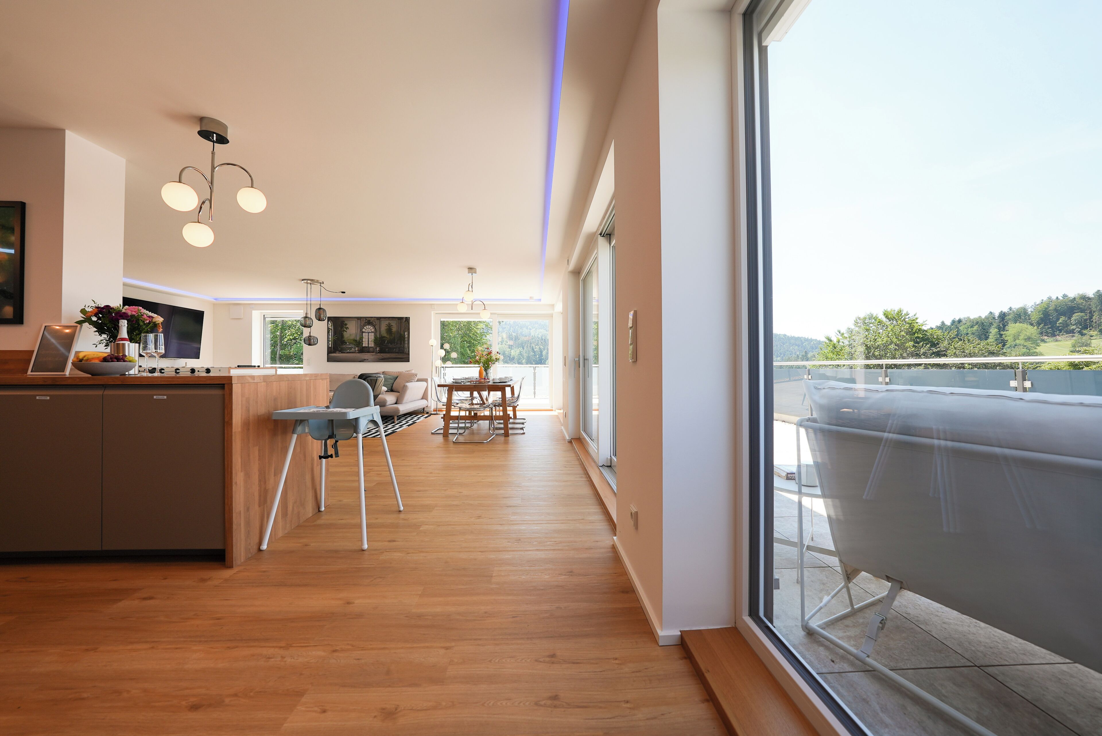 A modern kitchen with wooden flooring, a dining area with a table and chairs, and a large window with a view of a balcony.