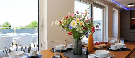 A dining table set with white dishes, glasses, and a vase of flowers, with a view of the outdoors through large windows.