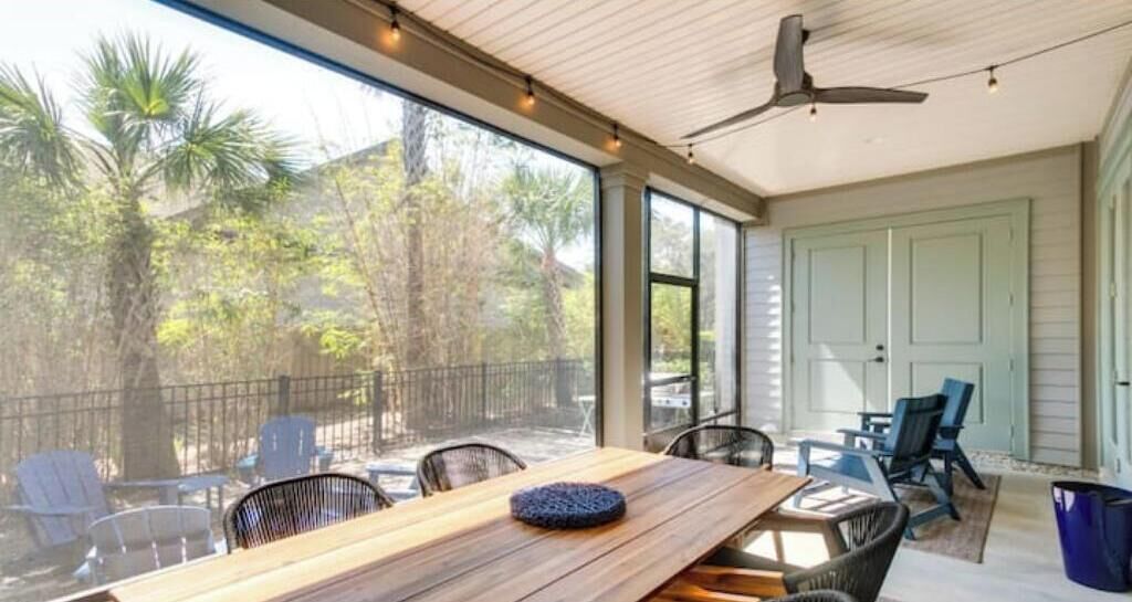 Une terrasse couverte avec une table et des chaises en bois, un ventilateur de plafond et une vue sur les arbres et une clôture.