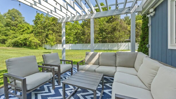 Patio esterno con una pergola bianca, divano beige e sedie di legno.