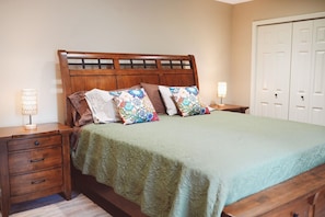 Un lit avec une tête de lit en bois, une couvre-lit verte et plusieurs oreillers. On y voit deux tables de chevet avec des lampes et une commode en bois avec des tiroirs. Une porte blanche est visible au fond.