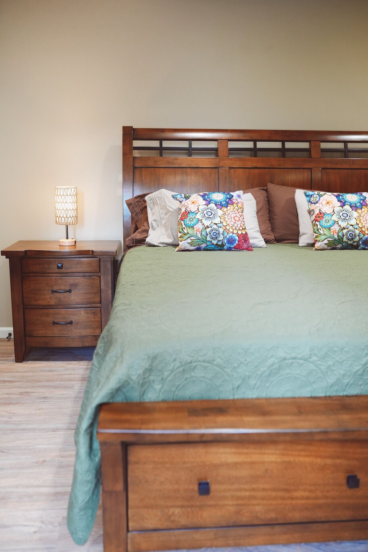 A bedroom with a wooden bed, green bedding, a nightstand with drawers, and a lamp.