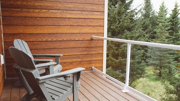 A wooden deck with Adirondack chairs, a glass balustrade, and a view of a forest.