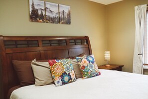 A bed with a wooden headboard, two patterned pillows, a nightstand with a lamp, and a window with curtains.