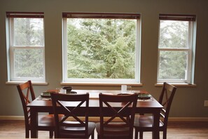 A dining area with a table and four chairs, three windows with a view of trees, and a light switch on the wall.