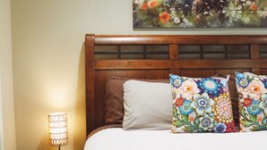 Habitació d'hotel amb un capçal de llit de fusta, un llit amb roba de llit blanca, dos coixins decoratius, una taula de nit amb una làmpada i un quadre floral sobre el llit.