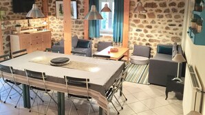 Un interior acogedor con pared de piedra, vigas de madera, una mesa de comedor con sillas, un sofá y una pequeña zona de cocina.