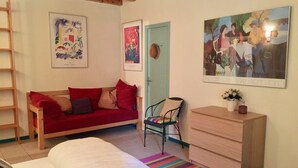 Una habitación con una cama, un sofá rojo, una silla, un aparador de madera y obras de arte en las paredes.