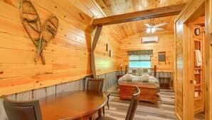 Interior de una cabaña de madera con comedor, cama y ventilador de techo.