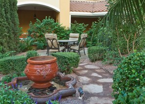 Une terrasse extérieure avec une table ronde et des chaises, entourée de haies soigneusement taillées et d’un grand pot en terre cuite.