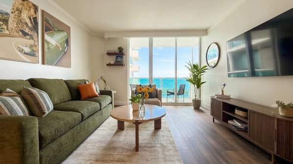 A living room with a green sofa, a coffee table, a TV, and a balcony with a view of the ocean.