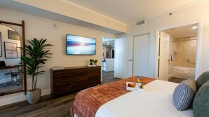 A modern hotel room with a large bed, a TV, a desk, a potted plant, and a shower area.