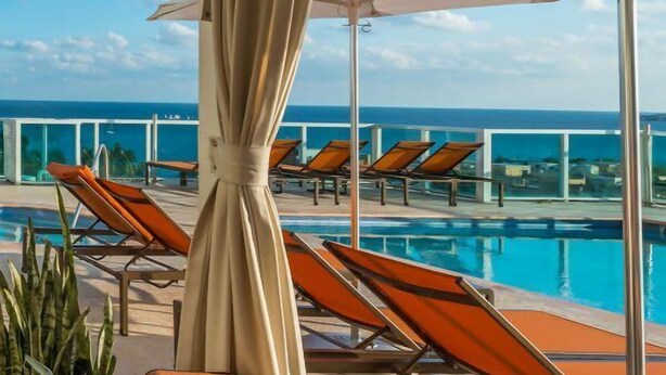 Un área junto a la piscina con sillones y sombrillas, con vista a un cielo azul despejado y al océano.