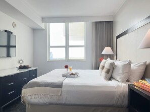 A hotel room with a large bed, a bedside table, a nightstand, a window with curtains, and a wall-mounted TV.
