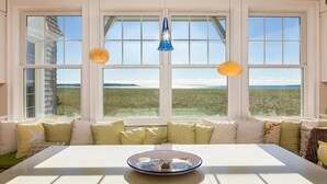 Une pièce dotée d’une grande fenêtre donnant sur un paysage verdoyant, une table blanche ornée d’un vase décoratif et un luminaire bleu suspendu au plafond.