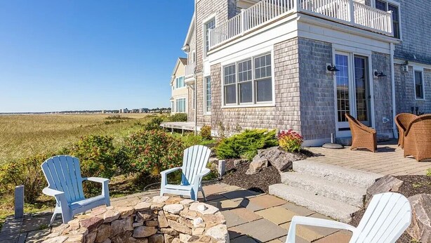 Une maison à deux étages dotée d’un balcon, d’un escalier en pierre et d’un espace extérieur aménagé avec des fauteuils Adirondack.
