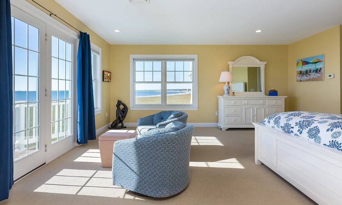 A bedroom with a bed, a chair, a dresser, and a large window with a view of the beach.