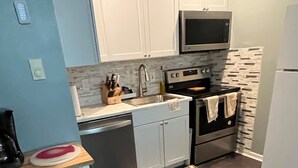 Una cocina moderna con armarios blancos, un lavamanos de acero inoxidable, un microondas y un horno. Hay una pequeña isla con un lavamanos y una cesta verde.