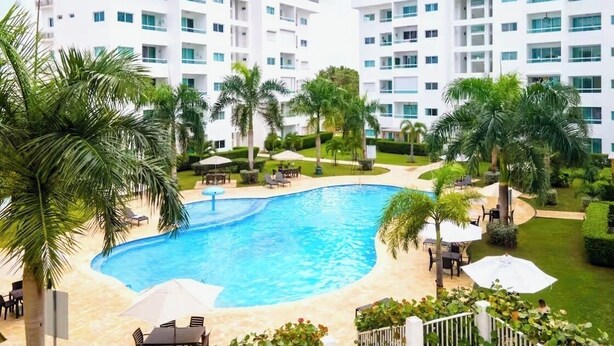A modern hotel with a central swimming pool surrounded by palm trees and outdoor seating areas. - Charming, elegant 2-bedroom condo with WiFi, AC in delightful Playa Juan Dolio (Playa Juan Dolio)