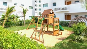 A playground with a swing set and slide, surrounded by greenery and a white building with balconies. - Charming, elegant 2-bedroom condo with WiFi, AC in delightful Playa Juan Dolio (Playa Juan Dolio)
