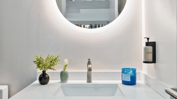 A modern bathroom with a round mirror, a white sink, a vase with green plants, a white candle, and a hand sanitizer dispenser. - High-Floor Residence in Coconut Grove (Miami)