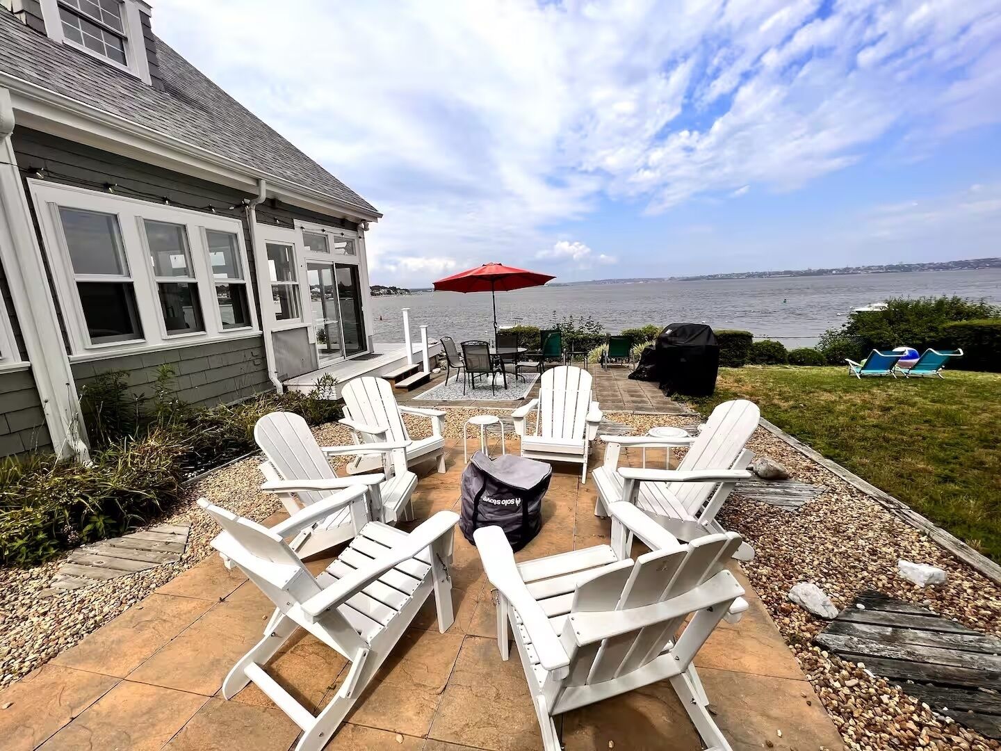 Eine Terrasse mit weißen Adirondack-Stühlen, einem roten Sonnenschirm und einem Grill.