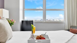 A hotel room with a bed, a tray with croissants, strawberries, and a glass of orange juice, and a view of the city through a large window.