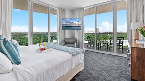 A hotel room with a large bed, a TV mounted on the wall, a balcony with a table and chairs, and a city view.